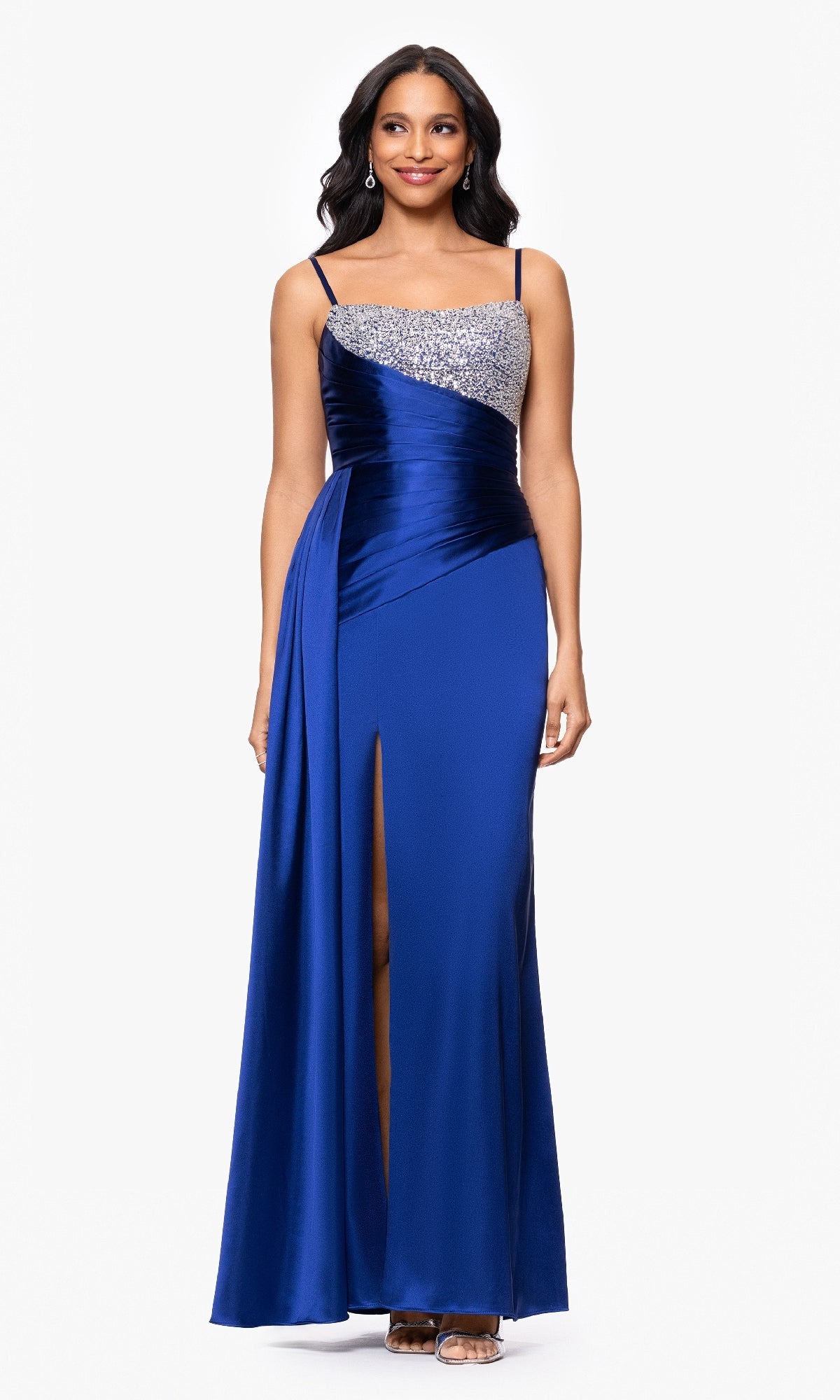 A woman wearing the Blondie Nites 5423BN long blue formal dress with a beaded bodice, thin straps, side drape, and front slit stands against a plain white background.